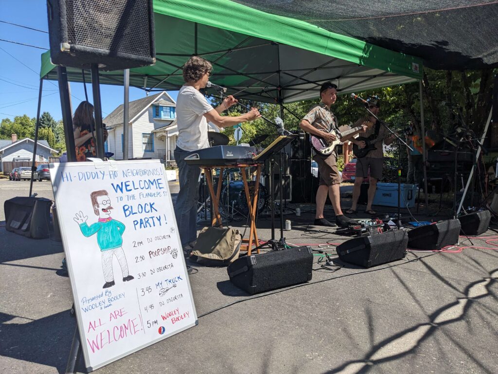 A live band performing under a green canopy tent at the Flanders Street Block Party, with a whiteboard sign in the foreground listing the day's schedule including performers DJ Oscurita, Poopship, My Truck, and Wooley Booley, and the message 'Hi-Diddly-Ho Neighborinos! All Are Welcome!'