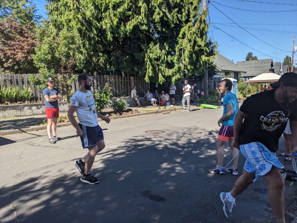 Several men playing a street game on a closed residential road on a sunny day, with spectators watching from the sidewalk and a cornhole board visible in the background.