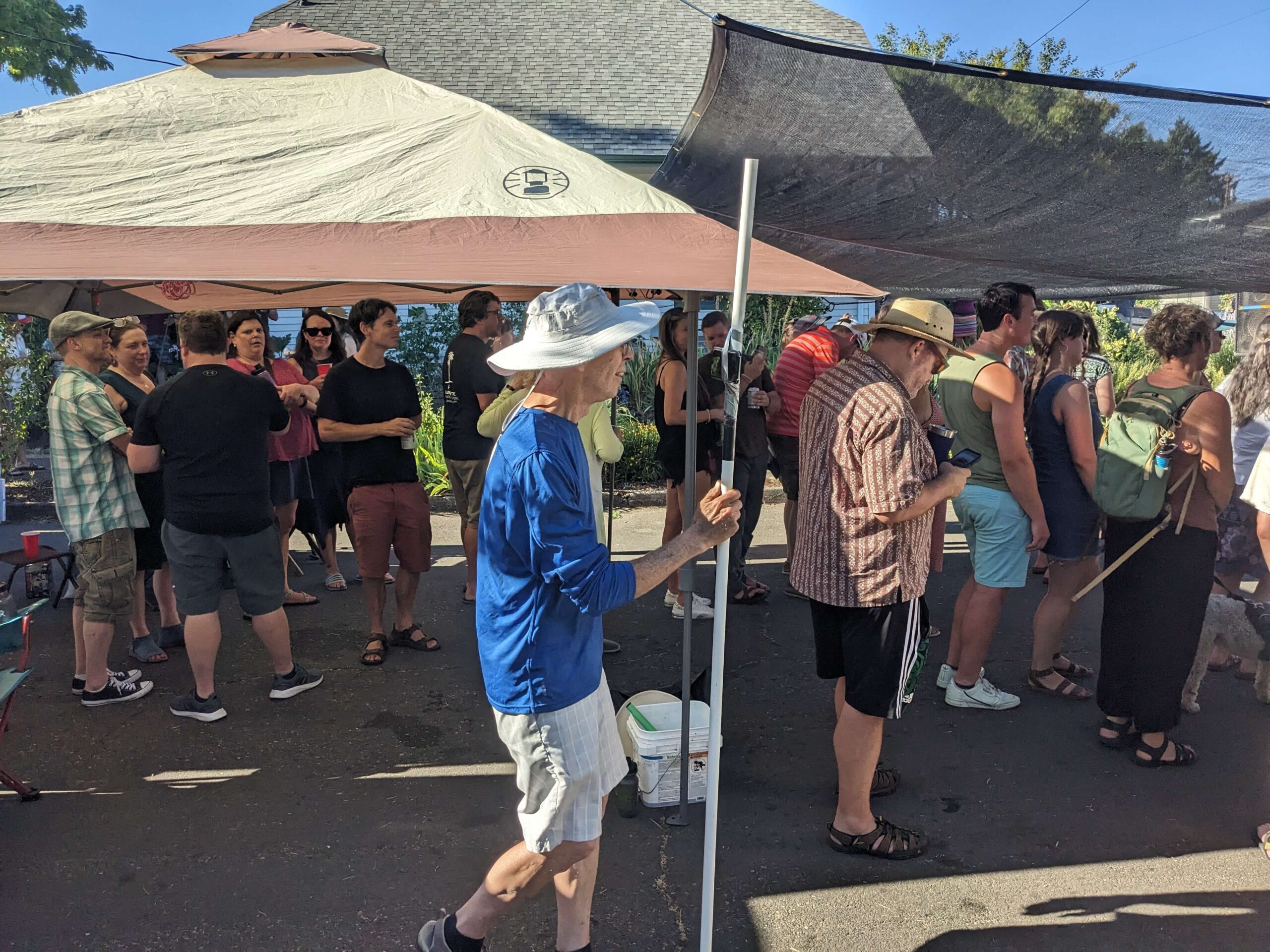 A crowd of adults socializing outdoors under pop-up canopy tents at a neighborhood block party on a sunny summer day, with an older man in a blue shirt and wide-brimmed sun hat walking in the foreground.