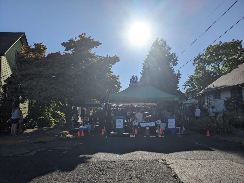 A sunny residential street blocked off for a neighborhood block party, with a green canopy tent, orange traffic cones, and handmade signs reading 'Street Closed,' 'All Are Welcome,' and 'Block Party,' surrounded by trees and houses.